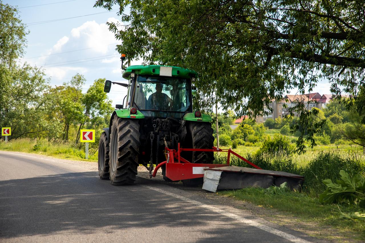 Big modern industrial tractor machine cutting green grass with mowing equipment along country roadside. Road lawn mower machinery vehicle highway maintenance service outdoors on sunny day.